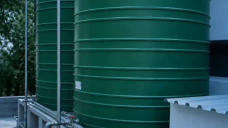 Two green vertical JoJo water storage tanks installed on elevated concrete plinths with neat galvanized steel inlet and outlet pipework.