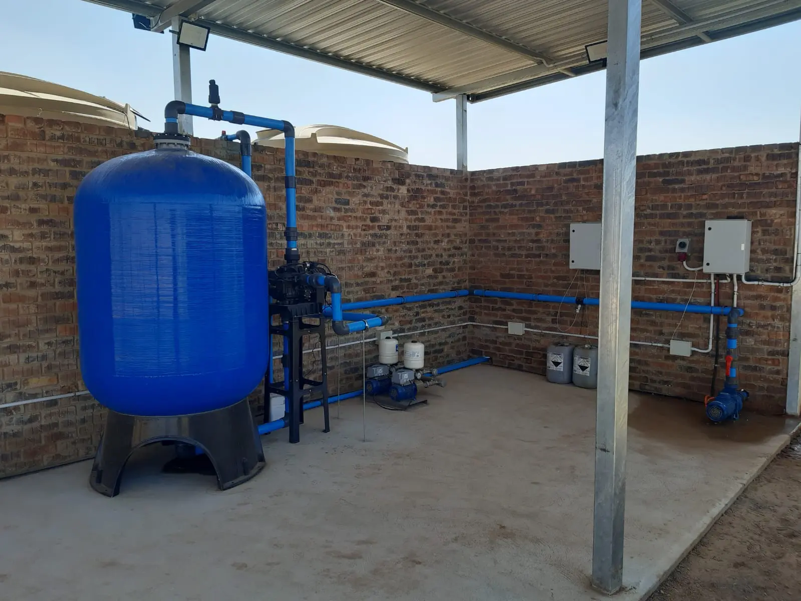 A commercial borehole filtration system under a canopy roof featuring a large blue FRP vessel with AFM glass media, a high-volume centrifugal pump, a dual booster pump set, and wall-mounted electrical control boxes.