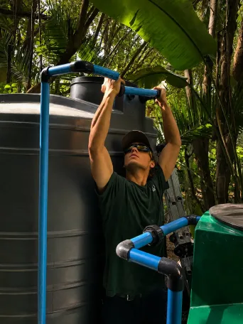 Technician fitting a blue inlet and overflow pipe assembly onto a large charcoal JoJo water storage tank.
